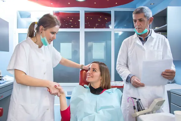 Female dentist with assistant shaking hands with female patient in the dentists chair.