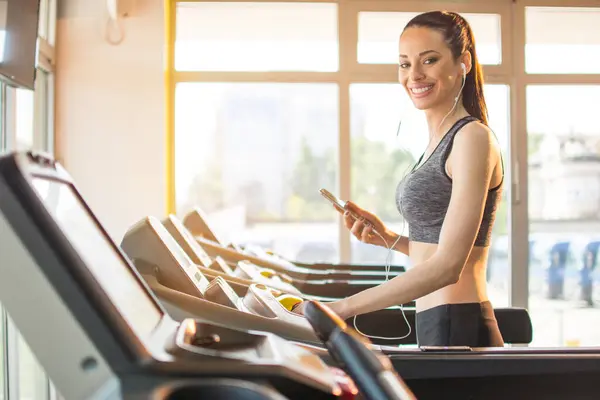 健身房的跑步机上戴着电话和耳机的健康的快乐女人 — 图库照片