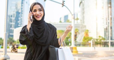 Arab woman in traditional wear holding shopping bags and talking on phone while walking on the street clipart