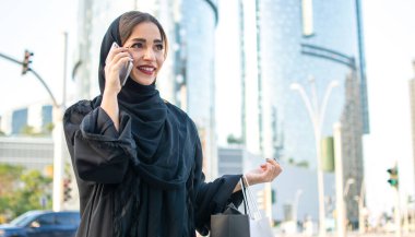 Arab woman in traditional wear holding shopping bags and talking on phone while walking on the street clipart