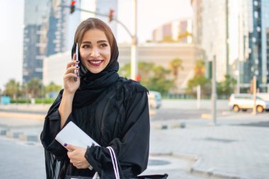 Geleneksel giysiler içinde, alışveriş çantaları ve cüzdan taşıyan ve sokakta cep telefonuyla konuşan güzel, genç Müslüman bir kadın..