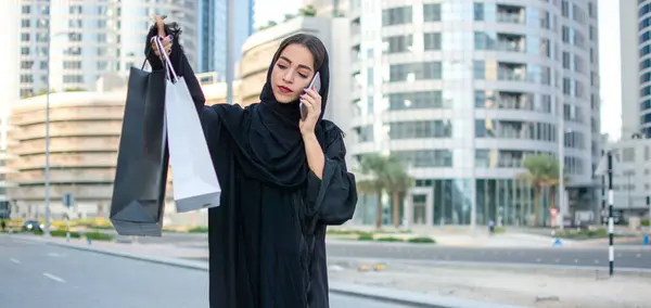 Arab woman in traditional wear holding shopping bags in hands and talking on mobile phone while walking on the city street
