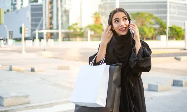 Arab woman in traditional wear holding shopping bags and talking on phone while walking on the street