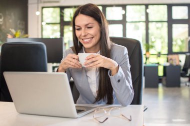Gülümseyen genç iş kadını ofiste dizüstü bilgisayar kullanırken kahve içiyor.
