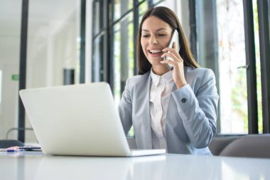 Gülümseyen iş kadını cep telefonuyla konuşuyor ve ofiste dizüstü bilgisayar kullanıyor.