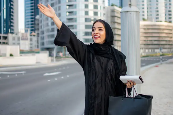 Young beautiful Arab woman in abaya with shopping bags, phone and wallet hitching a taxi on the street