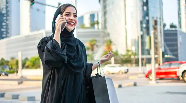 Arab woman in traditional wear holding shopping bags and talking on phone while standing on the street in front of the modern skyscrapers.