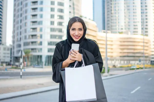 Excited Arab woman in traditional wear holding shopping bags and mobile phone while standing on the street in front of the modern skyscrapers.