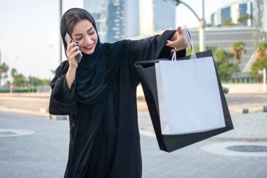 Arab woman in traditional wear holding shopping bags and talking on phone while standing on the street in front of the modern skyscrapers. clipart