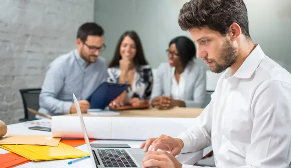 stock image Young handsome businessman working on laptop with coworkers discussing a new project in the background