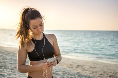 Kulaklıklı genç bir kadının plajdaki akıllı telefondan müzik ve mesaj dinlerken portresi.