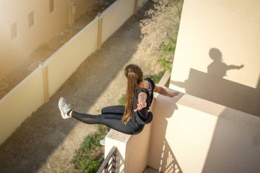 Neşeli kız manzarası açık havada duvarda oturuyor. Kaldırmış ellerle ve gün batımının keyfini çıkarıyor.