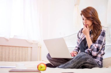 Genç bir kadın yatakta otururken dizüstü bilgisayar kullanıyor.