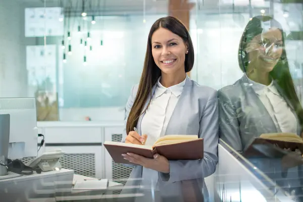 Young smiling business woman with notebook and pen standing in office and looking at camera