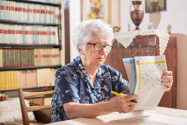 Старшая Женщина Сидит Столом Очках Чтения Концентрируясь Завершении Кроссворда Дома — стоковое фото