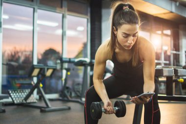 Athletic woman holding mobile phone and dumbbell while taking a break in gym clipart