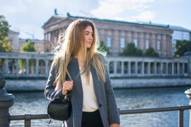 Güzel genç bir kadın, arka planda nehir ve sokak ile çitlere yaslanarak setin üzerinde dikilirken yüzünü çeviriyor. Rüzgardaki saçlar.