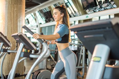 Spor salonunda eliptik antrenör makinesinde egzersiz yapan aktif genç bir kadın. Müzik dinleyen ve spor giyimli spor kıyafeti giyen eliptik antrenör makinesiyle spor yapan kız..