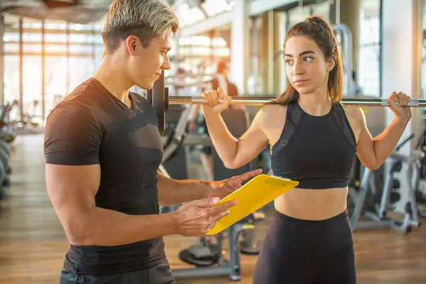 Male personal trainer and female client looking at progress date on clipboard at the gym. Young fit woman holding barbells and talking with personal trainer in gym.