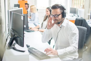 Kulaklık takmış genç bir adam parlak bir ofiste bilgisayar kullanıyor.