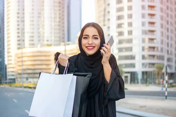 Cheerful young arab woman holding shopping bags and mobile phone on the street
