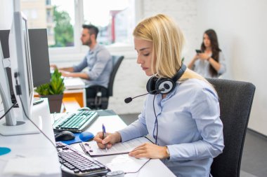 Kulaklık takmış güzel bir iş kadını ofis masasında iş belgelerini inceliyor.