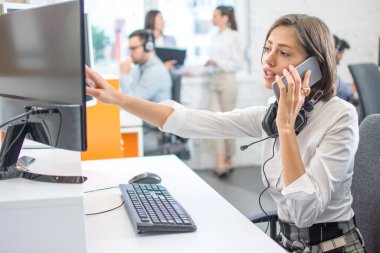 Güzel genç iş kadını ofiste çalışırken cep telefonuyla konuşuyor ve bilgisayardan bazı verileri kontrol ediyor.