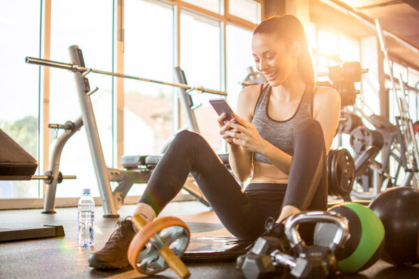Cheerful sporty girl using phone while sitting on mat in gym.
