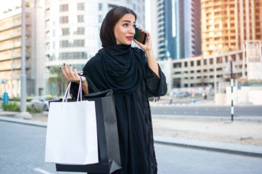 Arab woman in traditional wear holding shopping bags and talking on mobile phone while standing on the street in front of the skyscrapers clipart