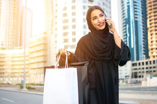 Arab woman in traditional wear holding shopping bags in hands and talking on mobile phone while walking on the street