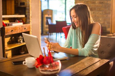Bir kafede cep telefonu ve laptop kullanan genç bir kadın.