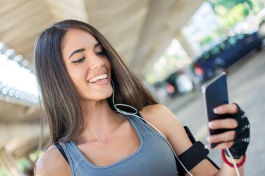 Genç, sportif bir kadın akıllı telefondan müzik dinliyor ve açık havada mesajlaşıyor..