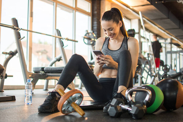 Beautiful fitness girl sitting and relaxing on exercise mat and using phone in the gym.