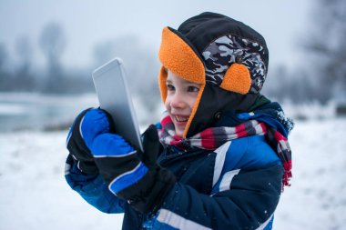 Karlı kış gününde akıllı telefonlu gülümseyen küçük bir çocuğun portresi.
