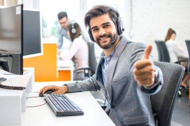 Dost canlısı erkek yardım hattı operatörü. Kulaklıklı adam çağrı merkezinde baş parmağını gösteriyor..