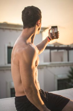 Genç kaslı adamın telefon tutuşu ve gün batımında dışarıda selfie çekişi..