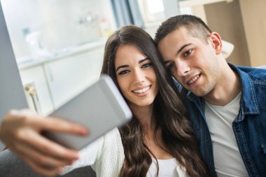 Genç, gülümseyen bir çift evde akıllı telefonuyla selfie çekiyor. Kapat.