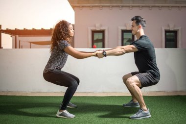Çatıdaki spor salonunda çömelme hareketleri yapan genç bir çift. Erkek ve kadın el ele tutuşup dışarıda çömelme egzersizi yapıyorlar..