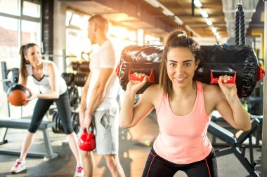 Genç, sportif bir kadın arka planda arkadaşlarıyla birlikte spor çantasını omuzlarına alıyor.