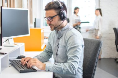 Ofisteki masaüstü bilgisayarında çalışan kulaklık takan yakışıklı erkek müşteri destek telefon operatörü.