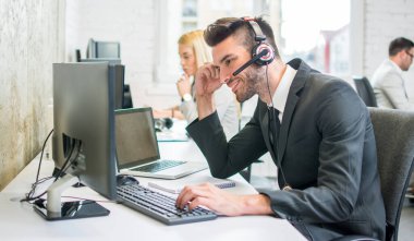 Ofiste bilgisayarla çalışan, kulaklık takan, gülümseyen bir erkek çağrı merkezi operatörü.