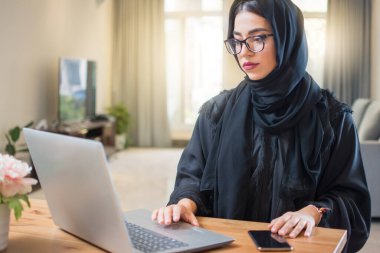 Evde dizüstü bilgisayarla çalışan tesettürlü genç bir Arap kadın..