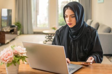 Evde bilgisayar ve akıllı telefon kullanan tesettürlü bir Arap kadın..