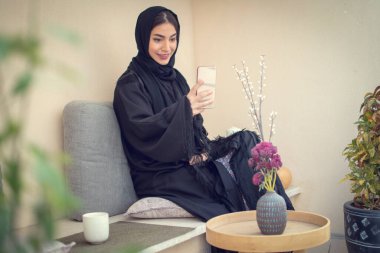 Güzel gülümseyen Arap kadın balkonda dinlenirken akıllı telefon kullanıyor..