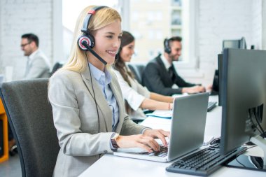 Gülümseyen arkadaş canlısı bayan müşteri destek operatörü. Kulaklıkla bilgisayar kullanıyor ve çağrı merkezinde müşteriyle konuşuyor.