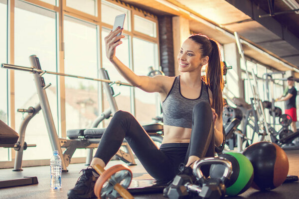 Beautiful girl in activewear using smart-phone to take a selfie photography during exercise break in gym.