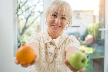 Evde kırmızı elma ve portakal tutan orta yaşlı ya da son sınıf kadını.