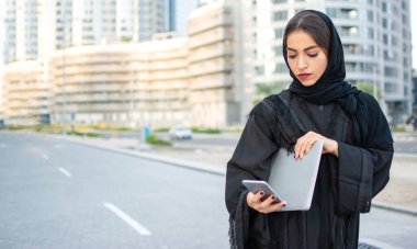 Güzel genç bir kadın dizüstü bilgisayar tutuyor ve sokakta telefon kullanıyor.