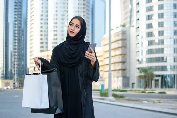 Arab woman in Abaya holding cell phone and shopping bags on the city street