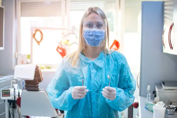 Female dentist in a protective suit with a protective shield and mask holding dental tools in dental office